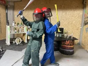 Two people in safety gear posing with a bat and crowbar in a rage room